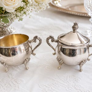 Puede incluir: Una lechera y un azucarero a juego con tapa, ambos en metal plateado. Las piezas presentan asas ornamentadas con volutas y tres patas decorativas. La lechera tiene un interior dorado y el azucarero incluye una tapa con un remate.