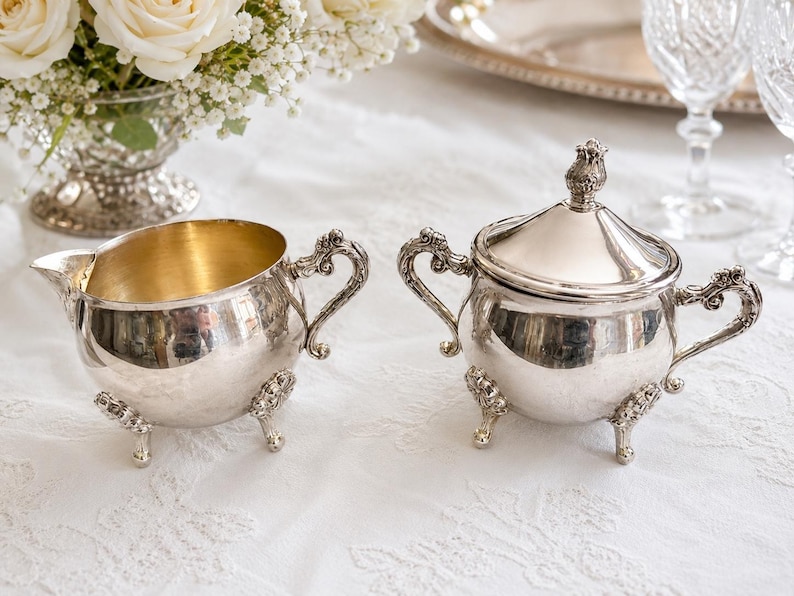Puede incluir: Una lechera y un azucarero a juego con tapa, ambos en metal plateado. Las piezas presentan asas ornamentadas con volutas y tres patas decorativas. La lechera tiene un interior dorado y el azucarero incluye una tapa con un remate.
