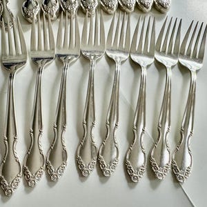 May include: A collection of silver forks with ornate floral detailing on the handles. The forks are arranged in rows, showcasing their elegant design. The tines are slender and the metal has a polished finish, reflecting light.