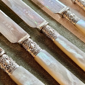 May include: Close-up of antique silverware. The knives have silver blades and ornate silver handles with a mother-of-pearl effect. The handles have a decorative, detailed design. The knives are arranged on a green and tan striped surface.