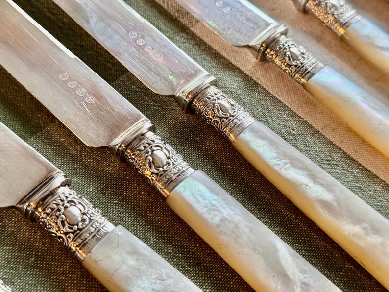 May include: Close-up of antique silverware. The knives have silver blades and ornate silver handles with a mother-of-pearl effect. The handles have a decorative, detailed design. The knives are arranged on a green and tan striped surface.
