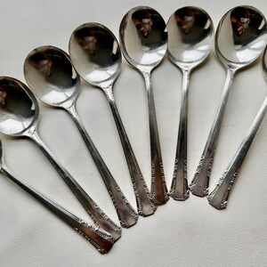May include: Set of eight silver-plated soup spoons with ornate handles. The spoons are arranged in a fan shape, with the bowls facing outwards.