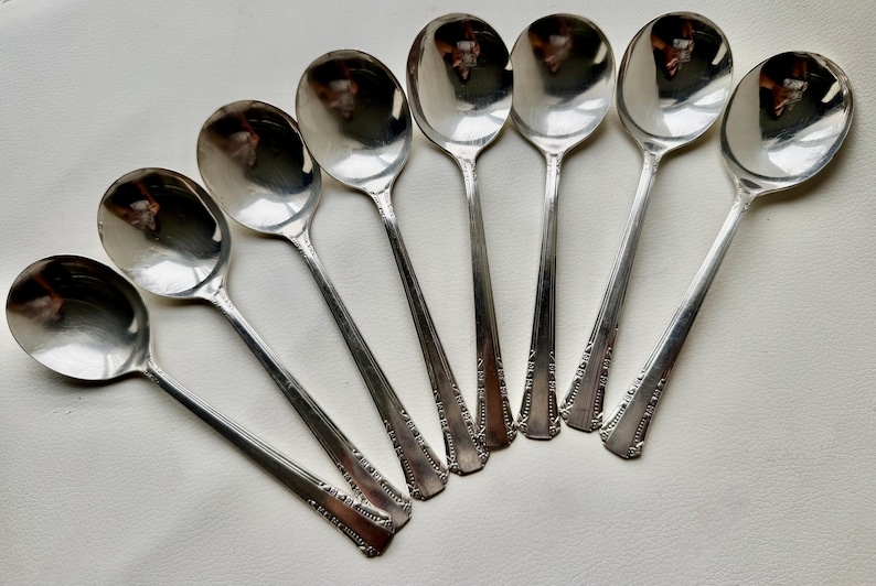 May include: Set of eight silver-plated soup spoons with ornate handles. The spoons are arranged in a fan shape, with the bowls facing outwards.