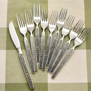 Puede incluir: Un juego de cubiertos de acero inoxidable que incluye un cuchillo de mesa y ocho tenedores. Cada pieza presenta un mango texturizado de color gris oscuro con un intrincado patr&oacute;n floral en relieve. Las cabezas son lisas, pulidas y plateadas.