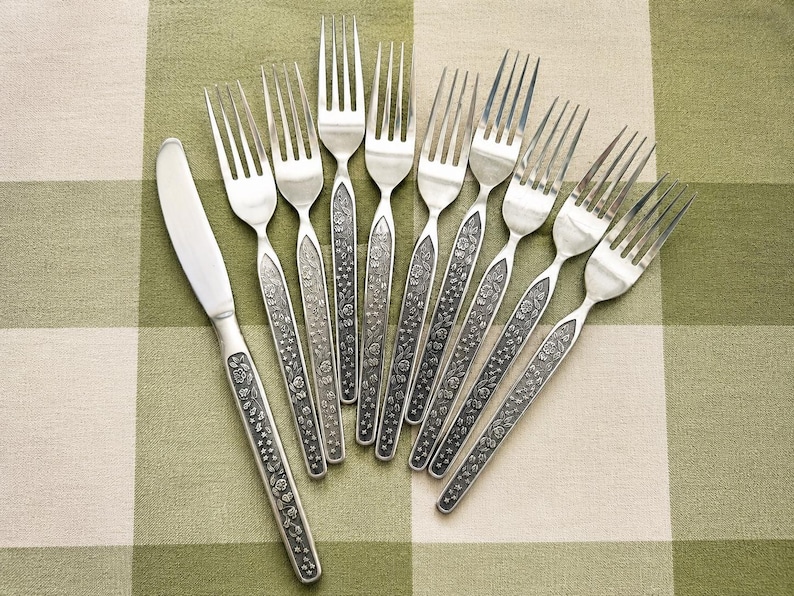 Puede incluir: Un juego de cubiertos de acero inoxidable que incluye un cuchillo de mesa y ocho tenedores. Cada pieza presenta un mango texturizado de color gris oscuro con un intrincado patr&oacute;n floral en relieve. Las cabezas son lisas, pulidas y plateadas.