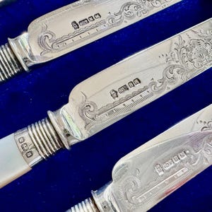 Może przedstawiać: Zestaw posrebrzanych noży z rękojeściami z opalizującej masy perłowej i żebrowanymi metalowymi skuwkami. Ostrza ozdobione są misternymi kwiatowymi grawerunkami i posiadają wyraźne cechy probiercze w pobliżu nasady ostrza.