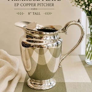 May include: A silver-toned EP copper pitcher with a curved handle and a wide pouring spout. The text above the pitcher reads VINTAGE PRIMROSE PLATE EP COPPER PITCHER 8" TALL. The pitcher has a polished, reflective surface and a circular base.