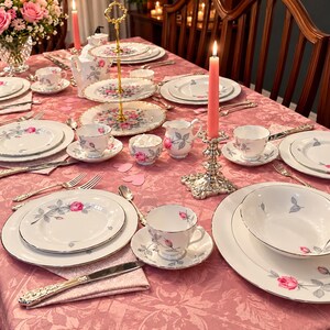 May include: A formal dining table setting featuring white porcelain dinnerware with a delicate pink rose and grey leaf pattern, silver-toned flatware, pink taper candles in ornate holders, and a tiered serving stand on a pink tablecloth.