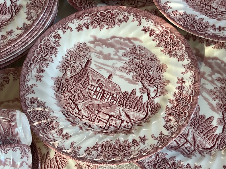 May include: A set of vintage pink and white floral patterned ceramic dinner plates. The plates feature a cottage scene with a red brick chimney and a red wooden fence.
