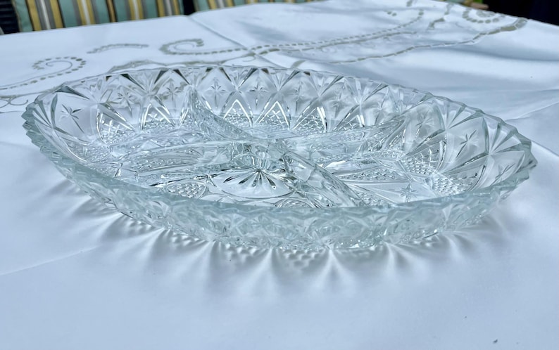 May include: Clear, oval-shaped glass serving dish with three compartments. The dish features a cut-glass design with star and diamond patterns. The edges are scalloped, and the dish sits on a white surface.