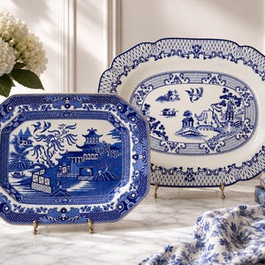 May include: Two ceramic platters featuring a blue and white willow pattern. The front rectangular platter displays a detailed landscape with pagodas and trees, while the larger oval platter behind it shows a similar scene with a scalloped border.