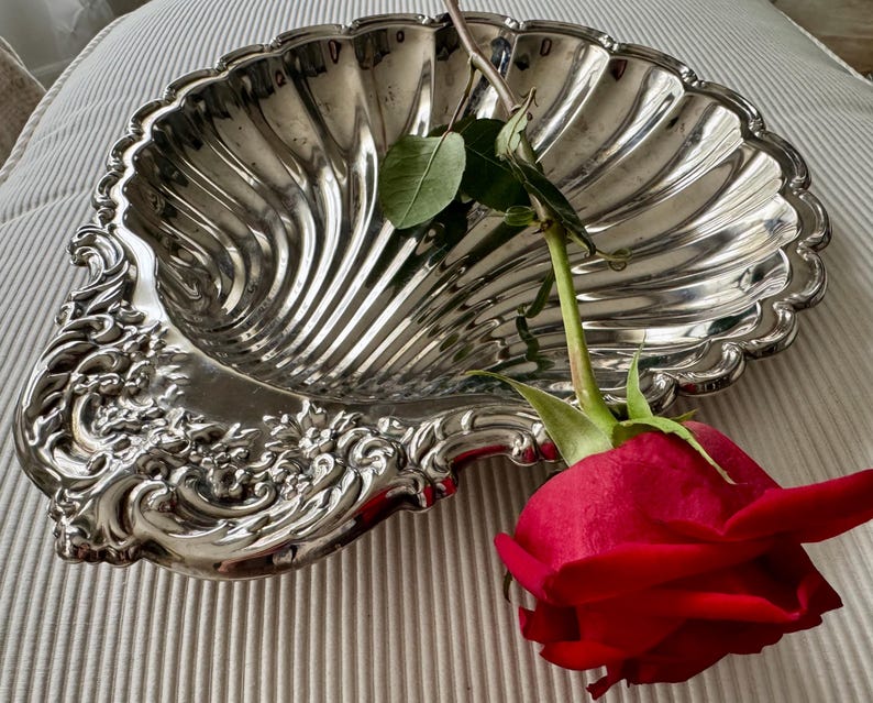 K&ouml;nnte beinhalten: Eine silberfarbene Servierschale in Form einer gro&szlig;en Jakobsmuschel mit gewelltem Rand und floraler Pr&auml;gung am Griffbereich. Eine einzelne rote Rose mit gr&uuml;nem Stiel und Bl&auml;ttern liegt diagonal &uuml;ber der Mitte der Schale.