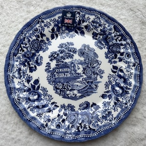 May include: A round ceramic plate featuring a blue and white transferware pattern. The center depicts a landscape with a house, bridge, and swans. The border is decorated with floral motifs. A Royal Wessex label with a crown and flag is attached.
