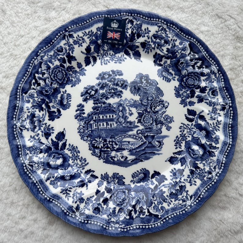 May include: A round ceramic plate featuring a blue and white transferware pattern. The center depicts a landscape with a house, bridge, and swans. The border is decorated with floral motifs. A Royal Wessex label with a crown and flag is attached.