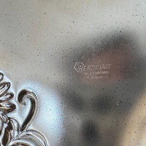 May include: Close-up of a silver-toned metal tray with embossed floral detailing. The tray has a textured surface and the text "REICHHART W. GERMANY SP. BRASS" is visible. The design features a flower and leaf pattern.