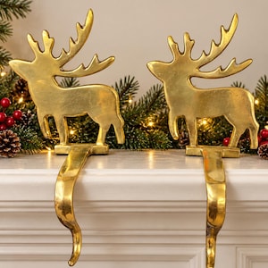 May include: Two gold-colored metal stocking holders, each featuring a standing reindeer silhouette with prominent antlers. The reindeer figures are attached to a curved hook designed to hang over the edge of a fireplace mantel.