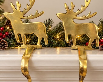 May include: Two gold-colored metal stocking holders, each featuring a standing reindeer silhouette with prominent antlers. The reindeer figures are attached to a curved hook designed to hang over the edge of a fireplace mantel.