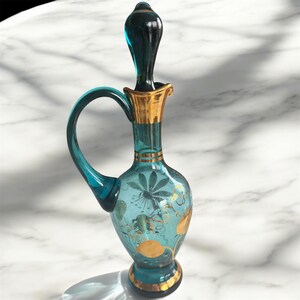 May include: A teal blue glass bottle with a gold stopper and intricate gold floral designs. The bottle has a handle and a round base.