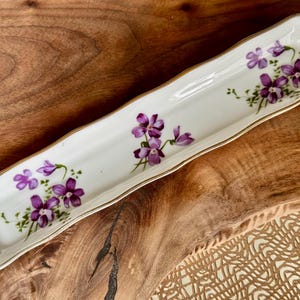 May include: A white ceramic trinket tray with a gold rim and a floral design featuring purple violets.