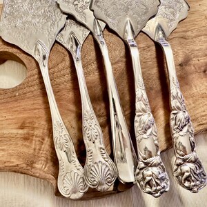 K&ouml;nnte beinhalten: Ein Set aus f&uuml;nf silberfarbenen Tortenhebern und Besteckteilen mit detaillierten, gepr&auml;gten Blumen- und Muschelmustern auf den Griffen und Klingen. Die Utensilien haben polierte, reflektierende Oberfl&auml;chen mit kunstvollen Gravuren.