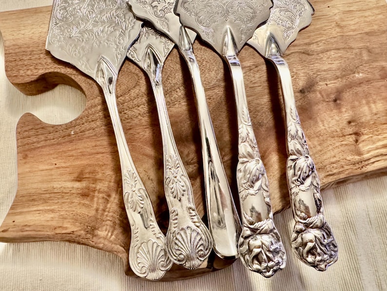K&ouml;nnte beinhalten: Ein Set aus f&uuml;nf silberfarbenen Tortenhebern und Besteckteilen mit detaillierten, gepr&auml;gten Blumen- und Muschelmustern auf den Griffen und Klingen. Die Utensilien haben polierte, reflektierende Oberfl&auml;chen mit kunstvollen Gravuren.