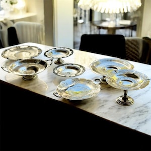 May include: A collection of antique silver serving dishes and trays, displayed on a marble surface. The pieces vary in size and design, including pedestal bowls and handled trays. The reflective surfaces catch the light, highlighting the intricate details and patterns.