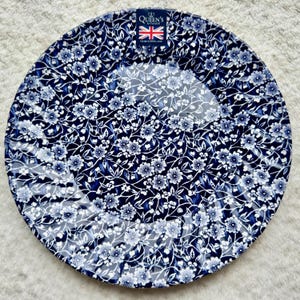 May include: A round ceramic plate featuring a dense, intricate white floral pattern set against a deep navy blue background. A rectangular label at the top displays the text Queen's by Churchill with a small Union Jack flag icon above the brand name.