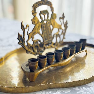 May include: A vintage brass menorah with seven candle holders, set on an oval tray. The menorah features a decorative crest with lions. The brass has a warm, aged patina, and the tray has handles.