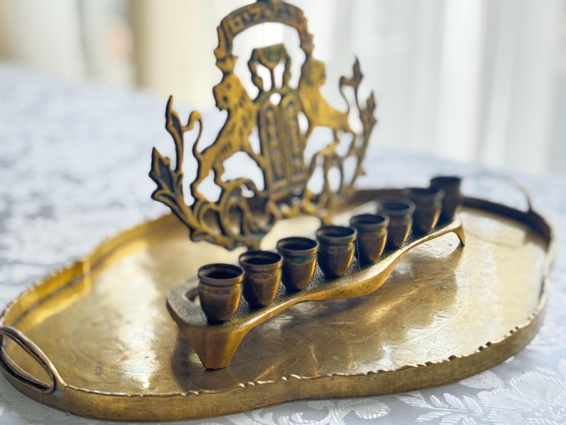 May include: A vintage brass menorah with seven candle holders, set on an oval tray. The menorah features a decorative crest with lions. The brass has a warm, aged patina, and the tray has handles.