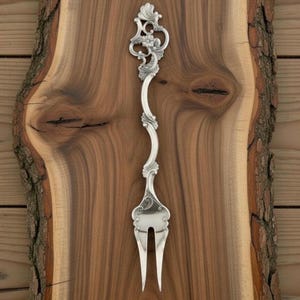 K&ouml;nnte beinhalten: Eine silberne Serviergabel mit einem verzierten, floralen Design. Der Griff ist anmutig geschwungen, und die Zinken sind schlicht und elegant. Die Gabel befindet sich vor einem h&ouml;lzernen Hintergrund.