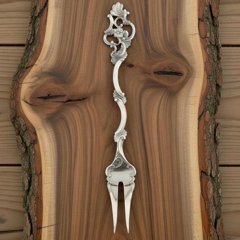 K&ouml;nnte beinhalten: Eine silberne Serviergabel mit einem verzierten, floralen Design. Der Griff ist anmutig geschwungen, und die Zinken sind schlicht und elegant. Die Gabel befindet sich vor einem h&ouml;lzernen Hintergrund.