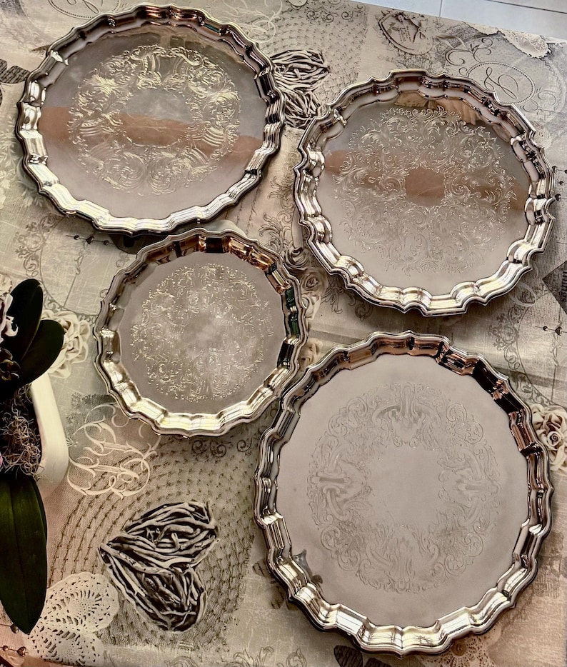 May include: A set of four round silver-toned metal trays with scalloped edges and intricate embossed floral patterns in the center. The trays vary in size and feature a reflective, polished finish on the surface and raised, curved rims.