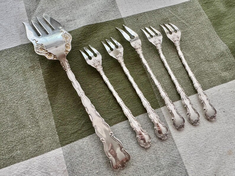 K&ouml;nnte beinhalten: Ein Set aus f&uuml;nf silberfarbenen Metallgabeln mit verzierten, gepr&auml;gten Blumendetails an den Griffen und am Hals. Die Kollektion umfasst eine gro&szlig;e Vorlegegabel und vier kleinere Gabeln, die alle eine polierte, reflektierende Metalloberfl&auml;che aufweisen.