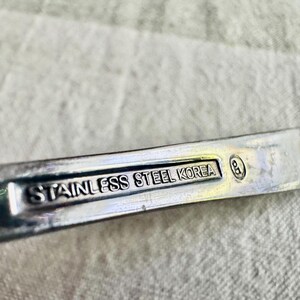 Może przedstawiać: Srebrzysty metalowy uchwyt przyboru kuchennego z wgłębionym prostokątnym panelem zawierającym wytłoczony napis STAINLESS STEEL KOREA oraz mały okrągły symbol cechy probierczej z literami GD umieszczony po prawej stronie napisu.