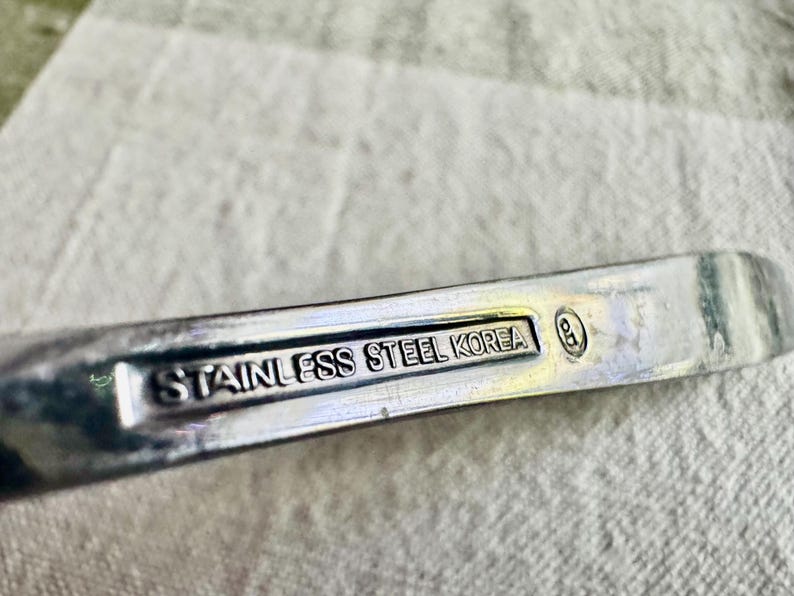 Może przedstawiać: Srebrzysty metalowy uchwyt przyboru kuchennego z wgłębionym prostokątnym panelem zawierającym wytłoczony napis STAINLESS STEEL KOREA oraz mały okrągły symbol cechy probierczej z literami GD umieszczony po prawej stronie napisu.