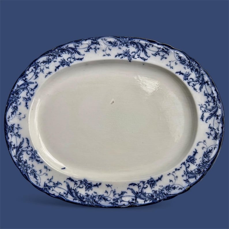 Puede incluir: Plato ovalado para servir con centro blanco y borde floral azul. El dise&ntilde;o azul presenta intrincados patrones de volutas y hojas. El plato tiene un borde festoneado y est&aacute; sobre un fondo azul.