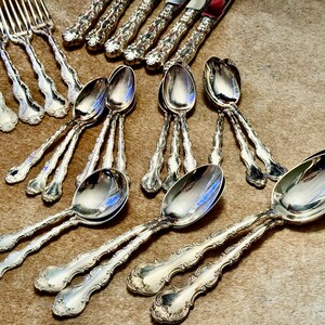 May include: A set of silver-plated silverware, including forks and spoons, with ornate designs. The silverware is arranged on a brown surface.