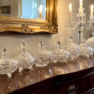 May include: A collection of seven clear cut glass lidded bowls with intricate geometric patterns and small feet. Each bowl features a matching lid with a pointed finial. The glass pieces are arranged in a row, showcasing reflective surfaces and detailed starburst motifs across the exterior of the bowls and lids.