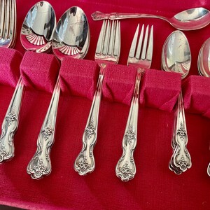 May include: A set of silver-toned flatware including forks and spoons with ornate floral detailing on the handles. The utensils are arranged in a storage case with a vibrant red velvet lining, featuring individual slots for each piece of cutlery.
