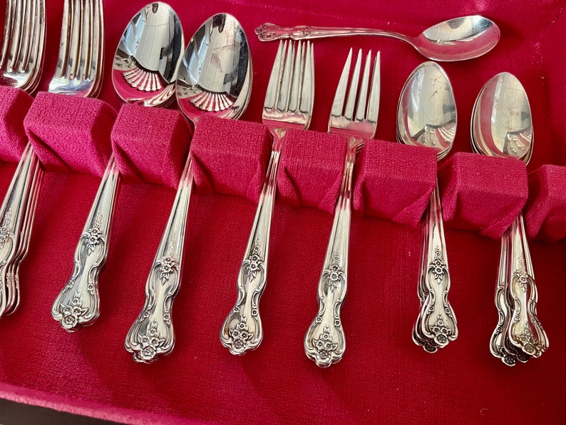 May include: A set of silver-toned flatware including forks and spoons with ornate floral detailing on the handles. The utensils are arranged in a storage case with a vibrant red velvet lining, featuring individual slots for each piece of cutlery.