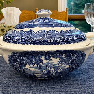 Op de afbeelding: Een witte keramieken terrine met een blauw transferware-patroon met een gedetailleerd landschap met een kasteel, bomen en gebladerte. Het ronde deksel heeft een bijpassende blauwe bloemenrand en een knop als handvat aan de bovenzijde.