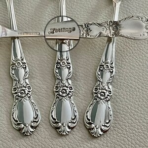 May include: Close-up of several silver spoons with ornate floral and scrollwork detailing. The spoons have a reflective surface and the word "Heritage" is visible on one of the spoon handles. The spoons are arranged on a light gray surface.