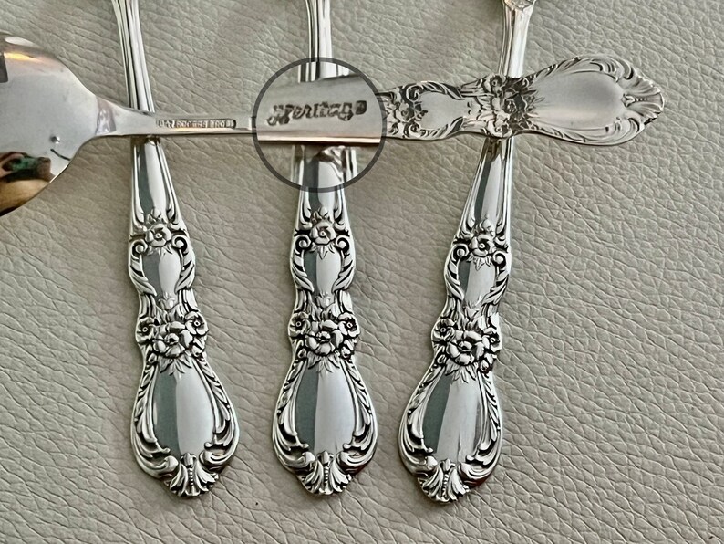 May include: Close-up of several silver spoons with ornate floral and scrollwork detailing. The spoons have a reflective surface and the word "Heritage" is visible on one of the spoon handles. The spoons are arranged on a light gray surface.