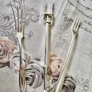 May include: Three silver serving utensils are displayed on a floral patterned cloth. The utensils include two forks and a cake lifter. The forks have different handle designs, and the cake lifter has a long handle with a clamp.