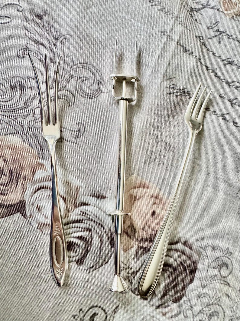 May include: Three silver serving utensils are displayed on a floral patterned cloth. The utensils include two forks and a cake lifter. The forks have different handle designs, and the cake lifter has a long handle with a clamp.