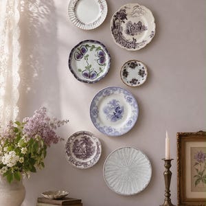 May include: A collection of seven decorative ceramic plates arranged on a wall, featuring various patterns including floral motifs, landscape scenes, and textured designs in shades of white, purple, and blue, alongside a candle and books.