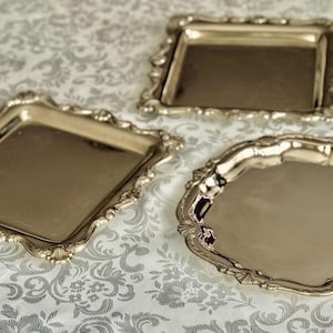 Puede incluir: Tres bandejas de metal plateado pulido con bordes festoneados ornamentados. Dos de las bandejas tienen forma cuadrada, mientras que la tercera bandeja es circular. Cada bandeja presenta una superficie lisa y reflectante con detalles decorativos.