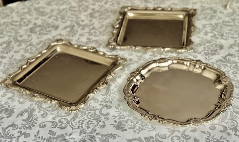 Puede incluir: Tres bandejas de metal plateado pulido con bordes festoneados ornamentados. Dos de las bandejas tienen forma cuadrada, mientras que la tercera bandeja es circular. Cada bandeja presenta una superficie lisa y reflectante con detalles decorativos.