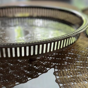 May include: A round silver-toned metal tray featuring a raised gallery edge with vertical slats and a beaded rim. The center of the tray displays an engraved floral pattern. The tray is supported by small, short feet and has a polished finish.