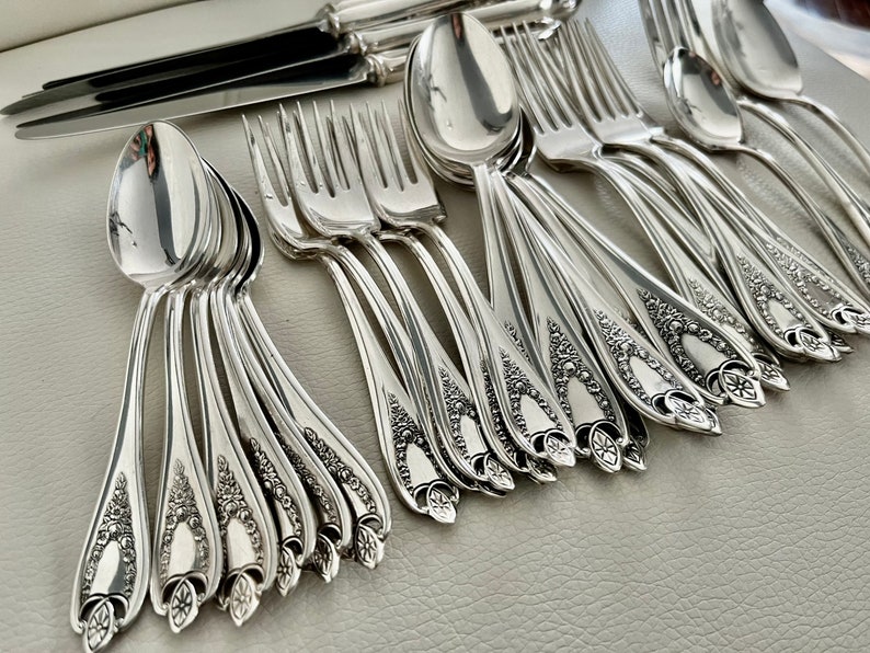 Puede incluir: Un conjunto de cubiertos plateados ornamentados, que incluye cucharas, tenedores y cuchillos. La cuberter&iacute;a presenta intrincados dise&ntilde;os florales en los mangos. El metal pulido refleja la luz, creando una exhibici&oacute;n brillante y elegante, ideal para una cena formal.